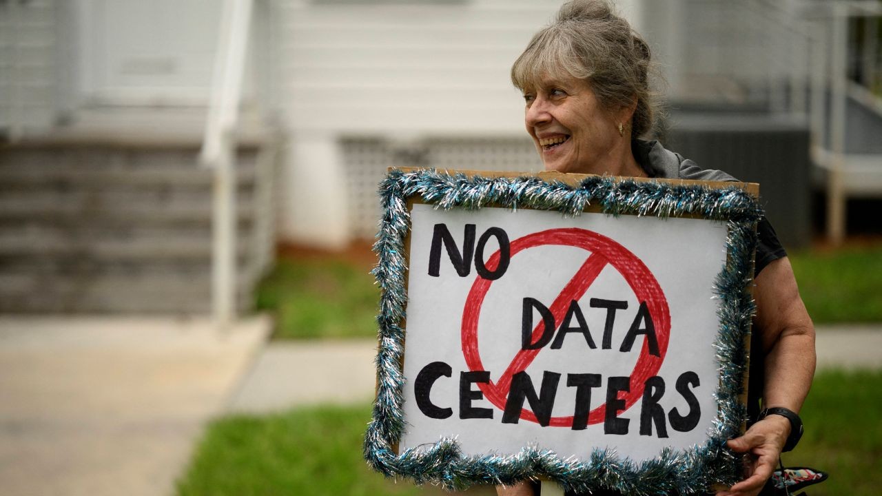 A smiling older woman holds a handmade sign outlined with tinsel that reads No Data Centers, with Data Centers crossed out by a red circle and slash. She stands outdoors in front of a light-colored building.