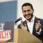 Abdul El-Sayed in a suit smiles and claps at a podium with a sign that reads FIGHT OLIGARCHY.