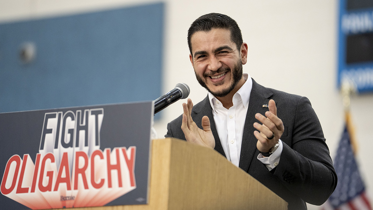 Home 5 Abdul El-Sayed in a suit smiles and claps at a podium with a sign that reads FIGHT OLIGARCHY.