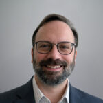 Smiling man with glasses, a beard, and mustache, wearing a blue blazer and collared shirt, standing in front of a plain light gray background.