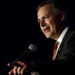A man in a suit and striped tie speaks into a microphone on a dark stage, gesturing with his hand.