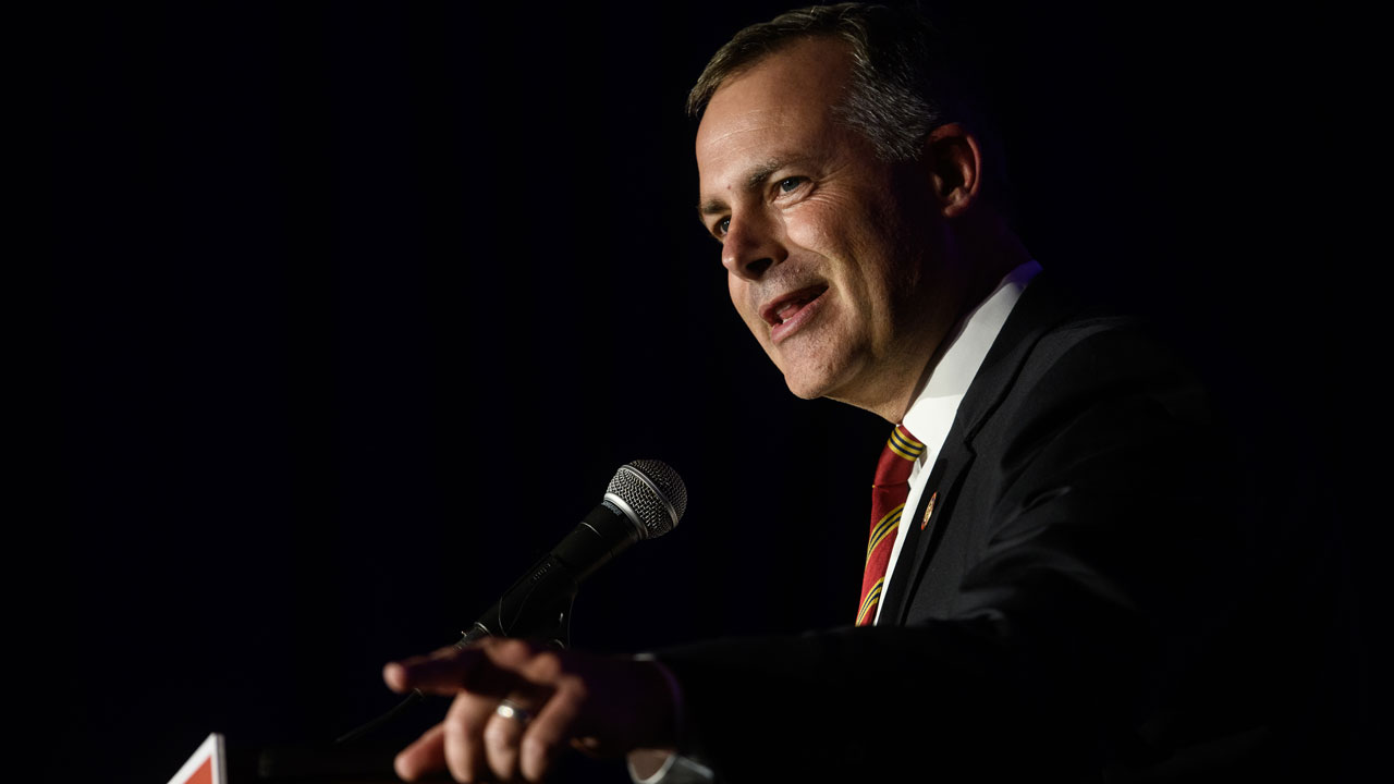 A man in a suit and striped tie speaks into a microphone on a dark stage, gesturing with his hand.