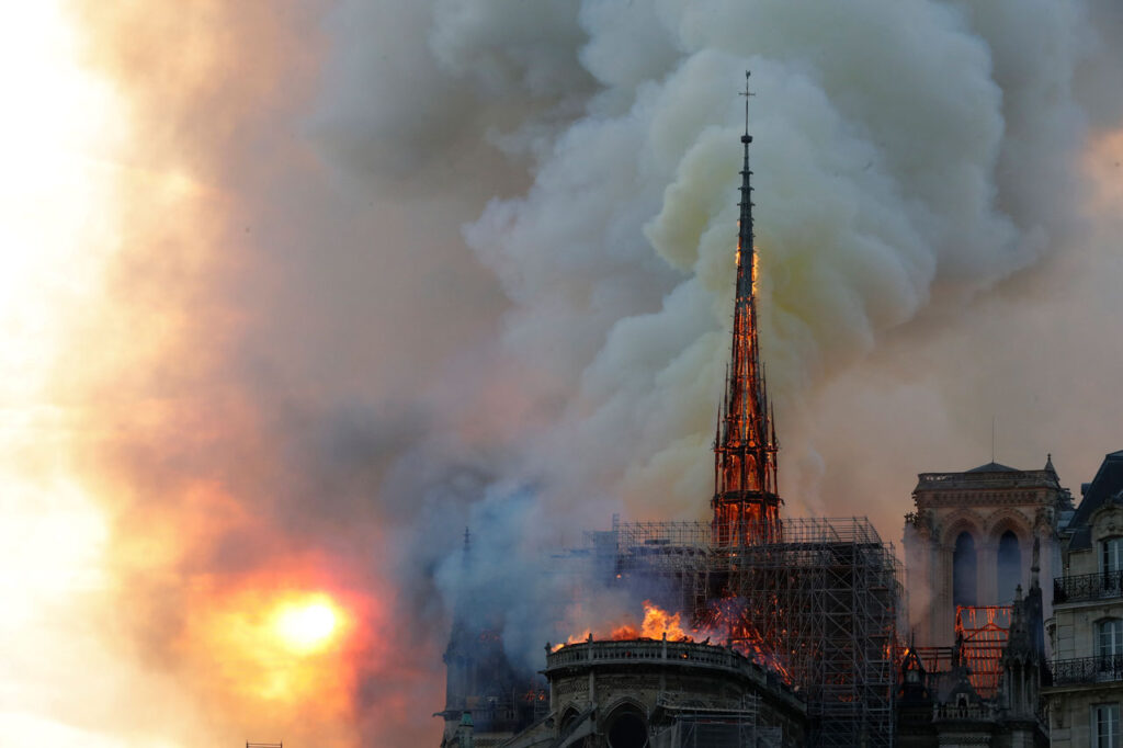 Bishop Barron’s First Theatrical Film Isn’t Nostalgia. It’s a Challenge. 3 Notre-Dame Cathedrals spire and roof engulfed in flames, with thick smoke billowing into the sky as the sun sets in the background.