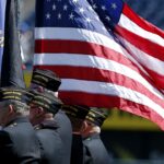 Three uniformed veterans stand in profile, carrying flags, including a large, unfurled American flag, during a ceremony at an outdoor event.
