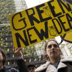 Two people stand outdoors holding a large yellow sign reading “Green New Deal” at a protest, with tall buildings and leafless trees visible in the background.