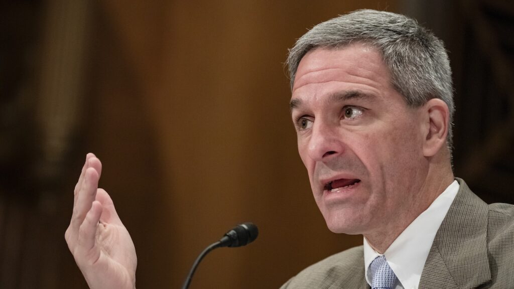 Ken Cuccinelli speaks into a microphone, gesturing with his left hand, appearing to address an audience or panel in a formal setting.