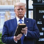 Trump holds up a closed book in front of a church sign outdoors. Shrubs and part of a building are visible in the background.