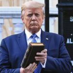 Trump holds up a closed book in front of a church sign outdoors. Shrubs and part of a building are visible in the background.