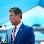 A man in a blue suit and red striped tie speaks at a podium with microphones, gesturing with his right hand, under a structure with blue geometric panels.