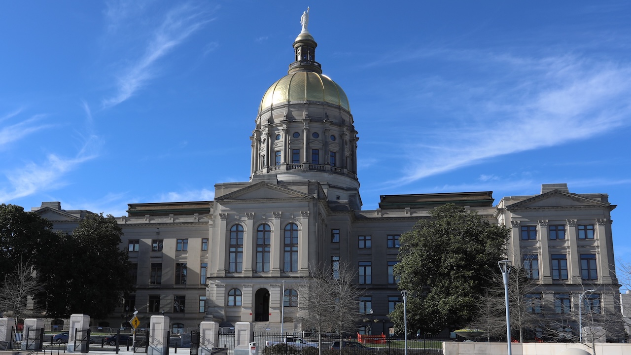 The Georgia State Capitol building sits with a gold dome and statue on top, set against a blue sky with wispy clouds. Trees and a fence are in front of the building.