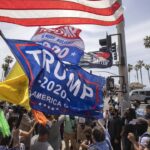 A crowd gathers outdoors by a street lined with palm trees, waving flags including a large Trump 2020 banner.