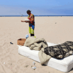 A person stands on a beach next to a pile of bedding and a box. There is a mattress, pillow, and blanket on the sand. Another person rides a bike in the background. The sky is clear and blue.
