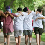 Six people walk arm-in-arm down a park path, facing away. One holds a small transgender pride flag.
