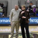 A man and a woman stand together on a basketball court, smiling. People are seated in the bleachers behind them, and a digital scoreboard displays the words Performance Therapy..