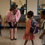 A woman in a pink jacket smiles and talks to three young children with backpacks in a school hallway. Another adult stands in the background, looking at a phone.