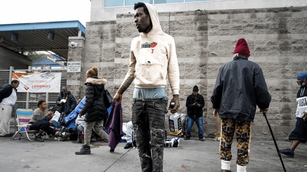 A young man in a hoodie and ripped jeans stands on a city street, looking to the side.