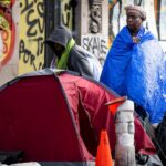 Two people stand near a red tent among belongings on a city street, wrapped in blankets, with graffiti on the wall behind them and bags and a traffic cone nearby.