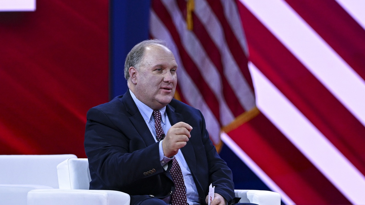 Home 5 A man in a dark suit and red tie sits on a white chair, gesturing with one hand. An American flag and red and white diagonal stripes are visible in the background.