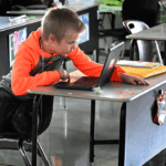 A young boy leans on his desk while using a laptop in a classroom. Papers and supplies are on the desk, and other empty desks are nearby.