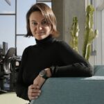 A person with short brown hair, wearing a black turtleneck and watch, sits on a light blue couch in a modern office with computers and a cactus in the background.
