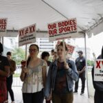 People hold signs reading OUR BODIES, OUR SPORTS and XY ≠ XX at an outdoor protest under a canopy. The group appears focused and engaged, standing together in an urban setting.