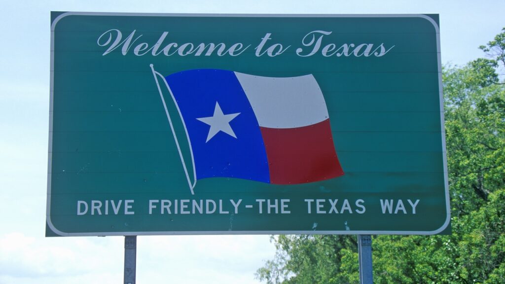 Green roadside sign with Welcome to Texas, a waving Texas flag, and the slogan Drive Friendly – The Texas Way against a backdrop of trees and blue sky.