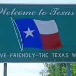 Green roadside sign with Welcome to Texas, a waving Texas flag, and the slogan Drive Friendly – The Texas Way against a backdrop of trees and blue sky.