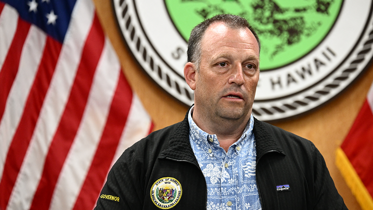 Home 2 A man in a blue patterned shirt and black jacket stands in front of Hawaii state and U.S. flags, speaking at a press event with an official seal visible in the background.