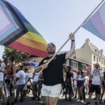 A group of people march in a parade, waving Progress Pride and transgender flags. The scene is sunny, and participants wear colorful clothes, celebrating LGBTQ+ pride on a city street lined with palm trees.