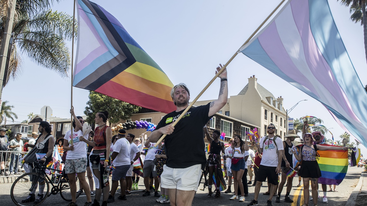 A group of people march in a parade, waving Progress Pride and transgender flags. The scene is sunny, and participants wear colorful clothes, celebrating LGBTQ+ pride on a city street lined with palm trees.