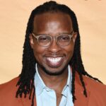A smiling man with glasses and long braided hair wearing a brown jacket and light blue shirt stands in front of a light tan background.