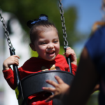 A young child smiles joyfully while swinging at a playground, wearing a red shirt and dark bow. An adult with a pink backpack stands nearby, gently pushing the swing. Trees and blurred playground equipment are in the background.