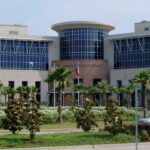 A modern courthouse building with large windows, surrounded by palm trees and greenery, with a few cars parked nearby and American and Texas flags at the entrance.