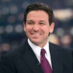 A man in a dark suit, white shirt, and purple tie smiles while seated against a blurred background with lights.