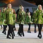 A group of women in matching green costumes perform an Irish dance on stage, smiling and stepping in unison. Bleachers and a tent are visible in the background.