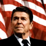 A man in a suit stands in front of several large, unfurled American flags, looking slightly upward with a serious expression.