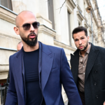 Two men in stylish suits walk outside near a stone building. One wears sunglasses and has a beard; photographers and others are visible around them. Trees with no leaves appear in the background.