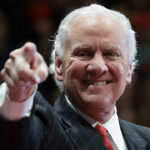 An older man in a suit and red tie smiles while pointing toward the camera. The background is blurred, focusing attention on his expression and gesture.