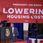 President Joe Biden speaks at a podium with a red sign behind him that reads Lowering Housing Costs during an event.