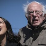 Two people stand outdoors against a blue sky; one is laughing with mouth wide open, the other, older, with white hair and glasses, looks forward with a neutral expression, both wearing jackets.