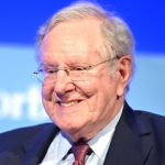 An older man in a suit and tie, embodying the confidence often associated with capitalism, smiles while sitting in front of a blue background with blurred white text.