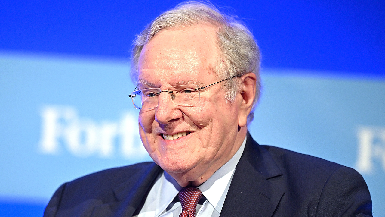 An older man in a suit and tie, embodying the confidence often associated with capitalism, smiles while sitting in front of a blue background with blurred white text.