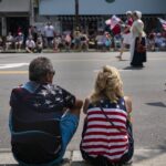 A man and woman, both wearing American flag-themed clothing, sit on the grass watching a parade with people carrying U.S. flags on the street. Spectators line the sidewalk in the background.