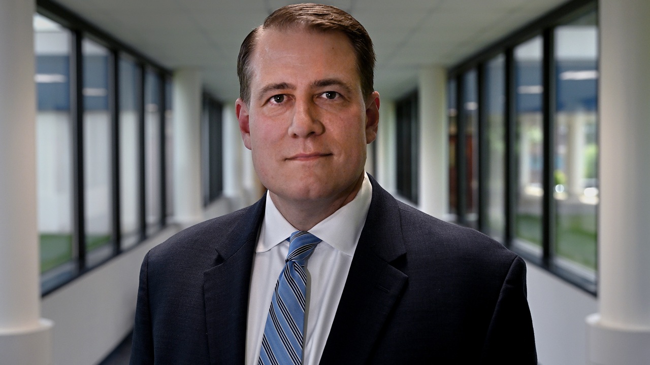 A man in a suit and striped tie stands in a bright hallway with large windows on both sides, looking directly at the camera.
