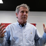 Sherrod Brown with gray hair in a light blue button-down shirt gestures with both hands while speaking indoors, standing in front of a whiteboard and red panel.