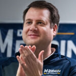 Republican Derek Merrin smiles as he applauds in front of one of his campaign signs.