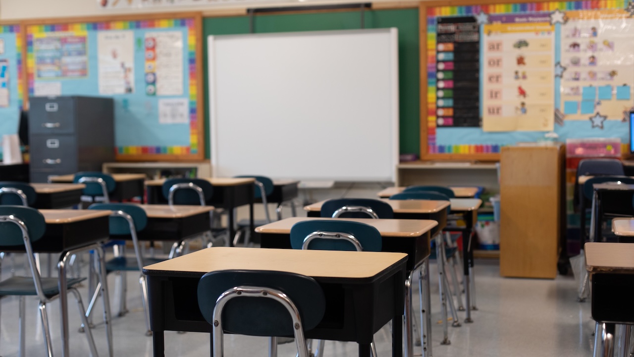 A classroom with empty desks and chairs faces a blank whiteboard. Colorful bulletin boards and educational posters decorate the walls. The room appears bright and organized.