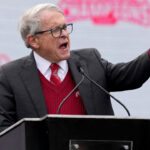 Gov. Mike DeWine gestures from the podium with an NCAA backdrop behind him.