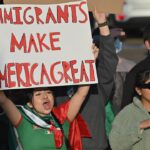 A woman in a crowd holds a sign that reads “Immigrants Make America Great.” Other people, including children and adults, stand around her, some clapping and wearing sunglasses or jackets.