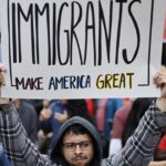 A person in a crowd holds a sign reading Immigrants make America great. People are gathered in the background, some holding flags, at an outdoor event or protest.